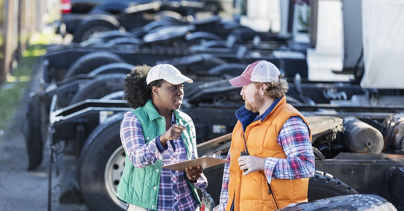 Multi-ethnic workers at a trucking company, standing by a fleet of semi-trucks, conversing. An African-American woman is holding a clipboard, giving directions to her coworker. She is in her 30s and the man is in his 40s.