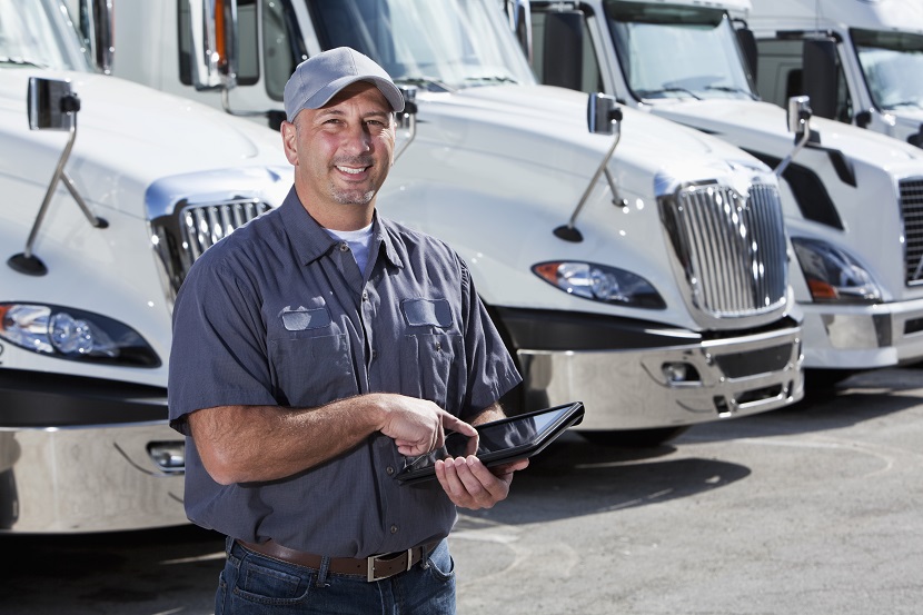 A truck driver standing in front of a row of parked semi-trucks, looking at the camera, holding a digital tablet. He is wearing a gray shirt, hat and blue jeans. He is smiling and confident. We see him from the waist up in the foreground of the image. He is a mid adult man, in his 30s.