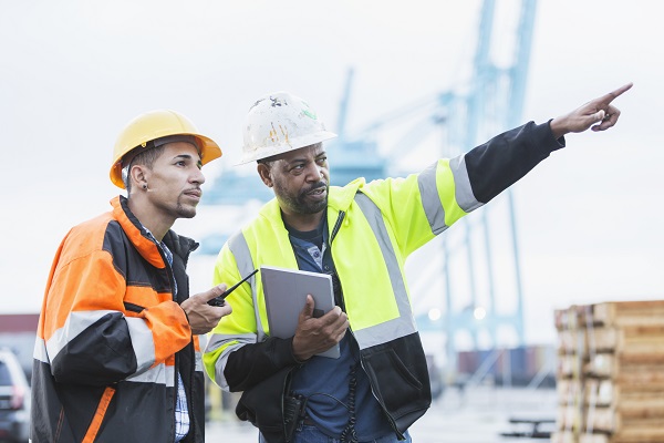 Two multi-ethnic men working at a shipping port. The mature African-American man in the yellow reflective jacket is in his 50s, holding a digital tablet and pointing in the distance. His mixed race Hispanic coworker is in his 30s.